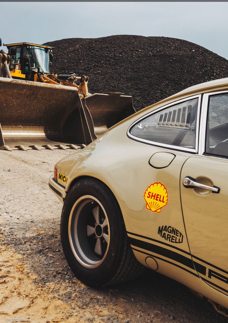 Vintage car with Shell and Magneti Marelli logos parked near a bulldozer in an industrial setting.
